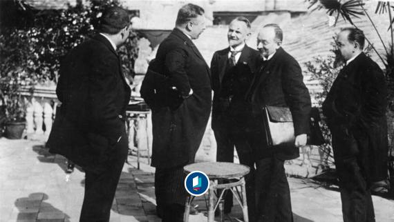 Schwarz-weiß Aufnahme von fünf Männer in Anzügen stehen im Freien und unterhalten sich in einer Gruppe.