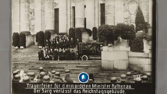 Trauerfeier mit vielen Menschen auf einer Treppe, ein Sarg wird aus einem Gebäude getragen, im Vordergrund mehrere Soldaten mit Helmen. Unten steht der Text: Trauerfeier für den ermordeten Minister Rathenau. Der Sarg verlässt das Reichtstagsgebäude.