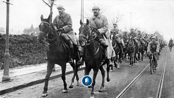 Schwarz-weiß-Fotografie einer Reiterstaffel in Uniform auf einer Straße mit Straßenbahnschienen, daneben ein Radfahrer.