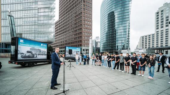 Ein Mann in einem Anzug spricht vor einer Gruppe von Jugendlichen, die vor einem Lastwagen am Potsdamer Platz steht. Darauf steht ein Spruch Sehnsucht nach Freiheit.