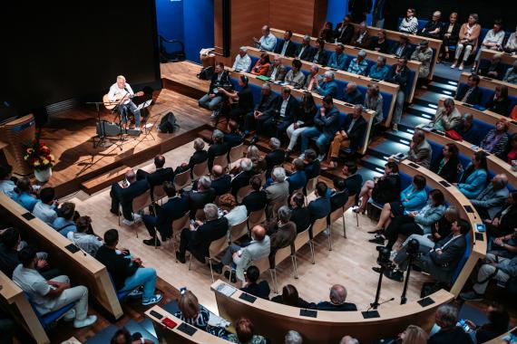 Ein Musiker spielt Gitarre auf einer Bühne vor einem Publikum in einem modernen, geschwungenen Saal mit Holzfußboden.