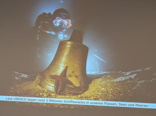 Ein Taucher mit einer Maske und Schnorchel schwebt über einer großen, goldfarbenen Glocke, die auf dem Meeresboden liegt. Ein Seestern ist auf der Glocke sichtbar.