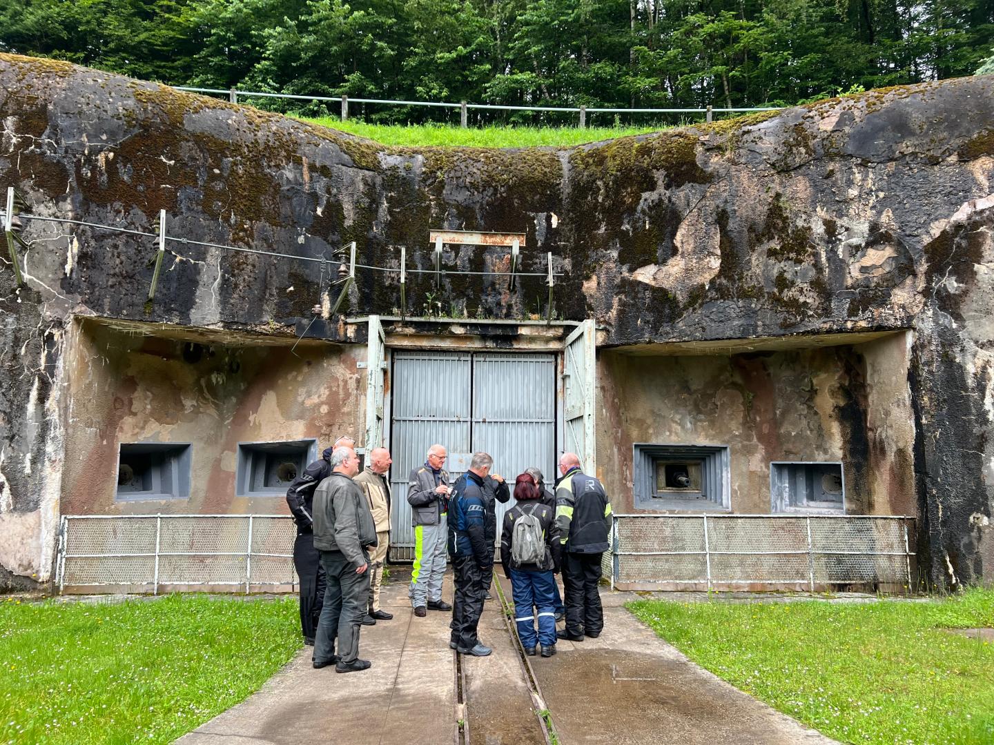 Sechs Personen stehen vor einem alten, verfallenen Bunker mit einer großen, grauen Metalltür und mehreren Fenstern. Die Wände sind von Moos und Verwitterung geprägt.