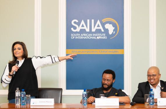 Eine Frau steht mit Mikrofon in der Hand und zeigt auf ein blaues Poster mit dem Schriftzug 'SAIIA SOUTH AFRICAN INSTITUTE OF INTERNATIONAL AFFAIRS African perspectives. Global insights.' Zwei Männer sitzen an einem Tisch mit Wasserflaschen davor.