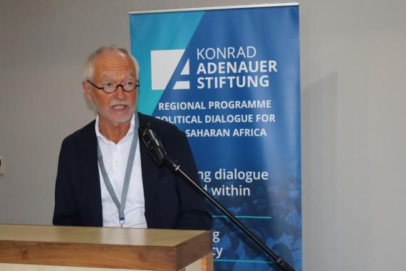 Mann mit weißem Hemd und dunklem Sakko steht an einem Rednerpult vor einem Banner der Konrad-Adenauer-Stiftung mit englischem Text