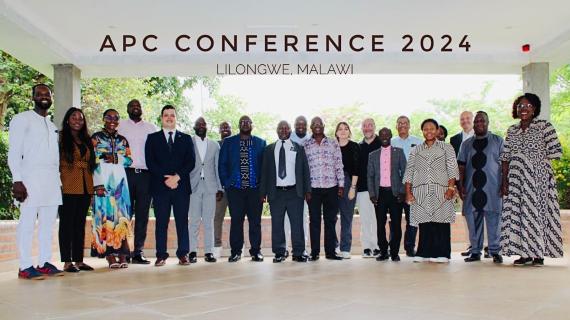 Gruppe von Menschen in formeller Kleidung steht unter einem überdachten Bereich mit der Aufschrift 'APC CONFERENCE 2024 LILONGWE, MALAWI' im Hintergrund