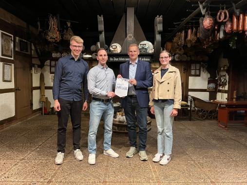 Vier Personen stehen in einem rustikalen Raum, zwei Männer in der Mitte halten ein weißes Stück Stoff mit der Aufschrift 'DORFLIEBE'