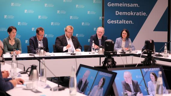 Demokratie gemeinsam gestalten steht auf einer Wand im Hintergrund. Expertinnen und Experten in einem Kongressraum mit Kameras und dem Logo der Konrad-Adenauer-Stiftung