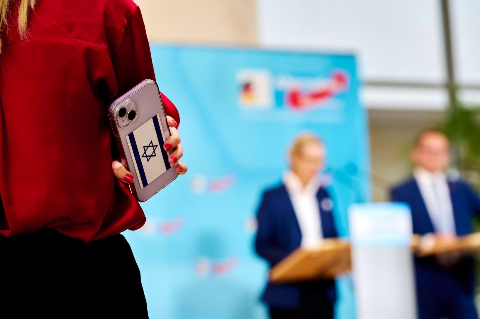 Handy mit Israel Flagge im Vordergrund, im Hintergrund Tino Chrupalla und Alice Weidel von der AfD bei einer Pressekonferenz