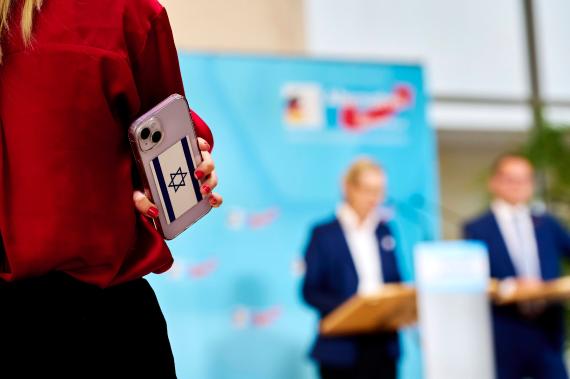 Handy mit Israel Flagge im Vordergrund, im Hintergrund Tino Chrupalla und Alice Weidel von der AfD bei einer Pressekonferenz