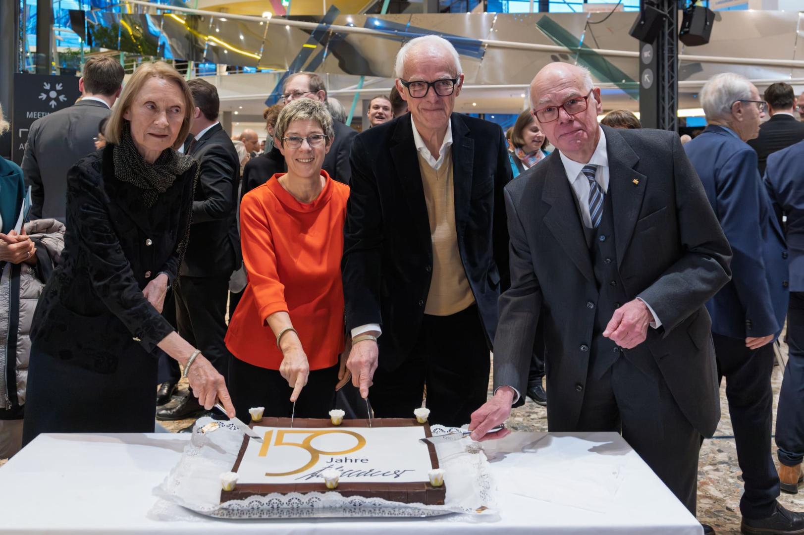 Vier Personen stehen an einem Tisch und schneiden gemeinsam eine rechteckige Torte mit der Aufschrift '150 Jahre' an.