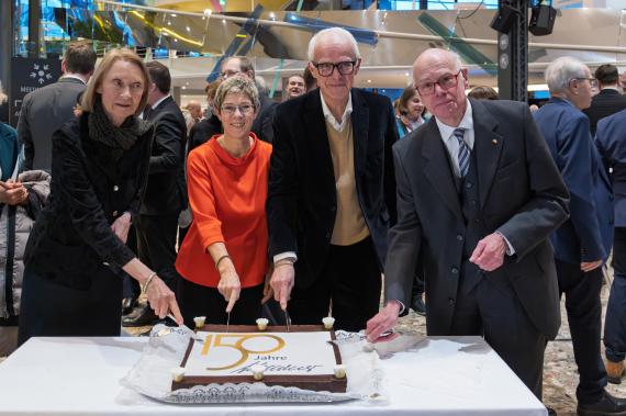 Vier Personen stehen an einem Tisch und schneiden gemeinsam eine rechteckige Torte mit der Aufschrift '150 Jahre' an.