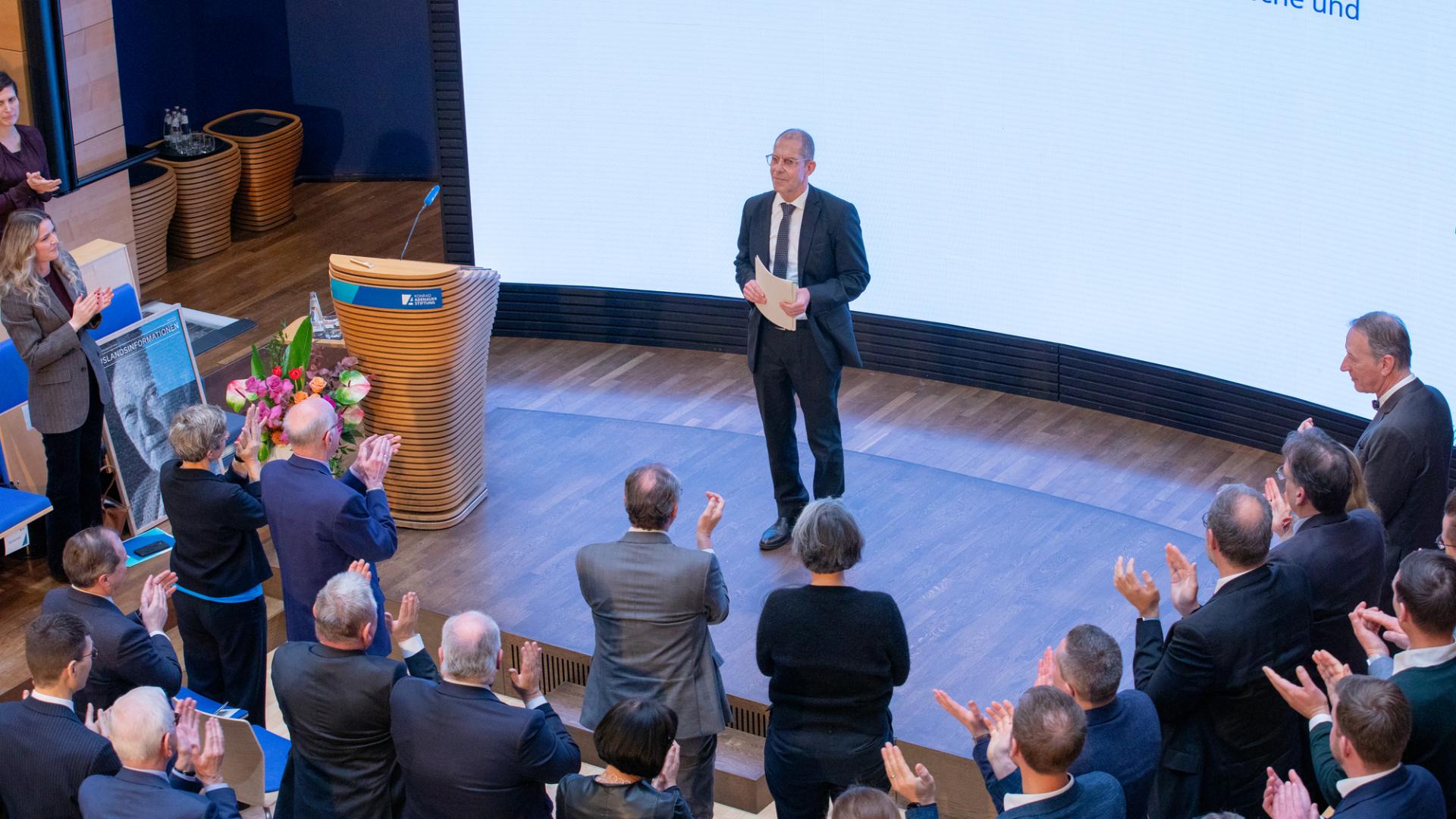 Ein Mann in Anzug hält eine Rede auf einer Bühne vor einem Publikum, das applaudiert.