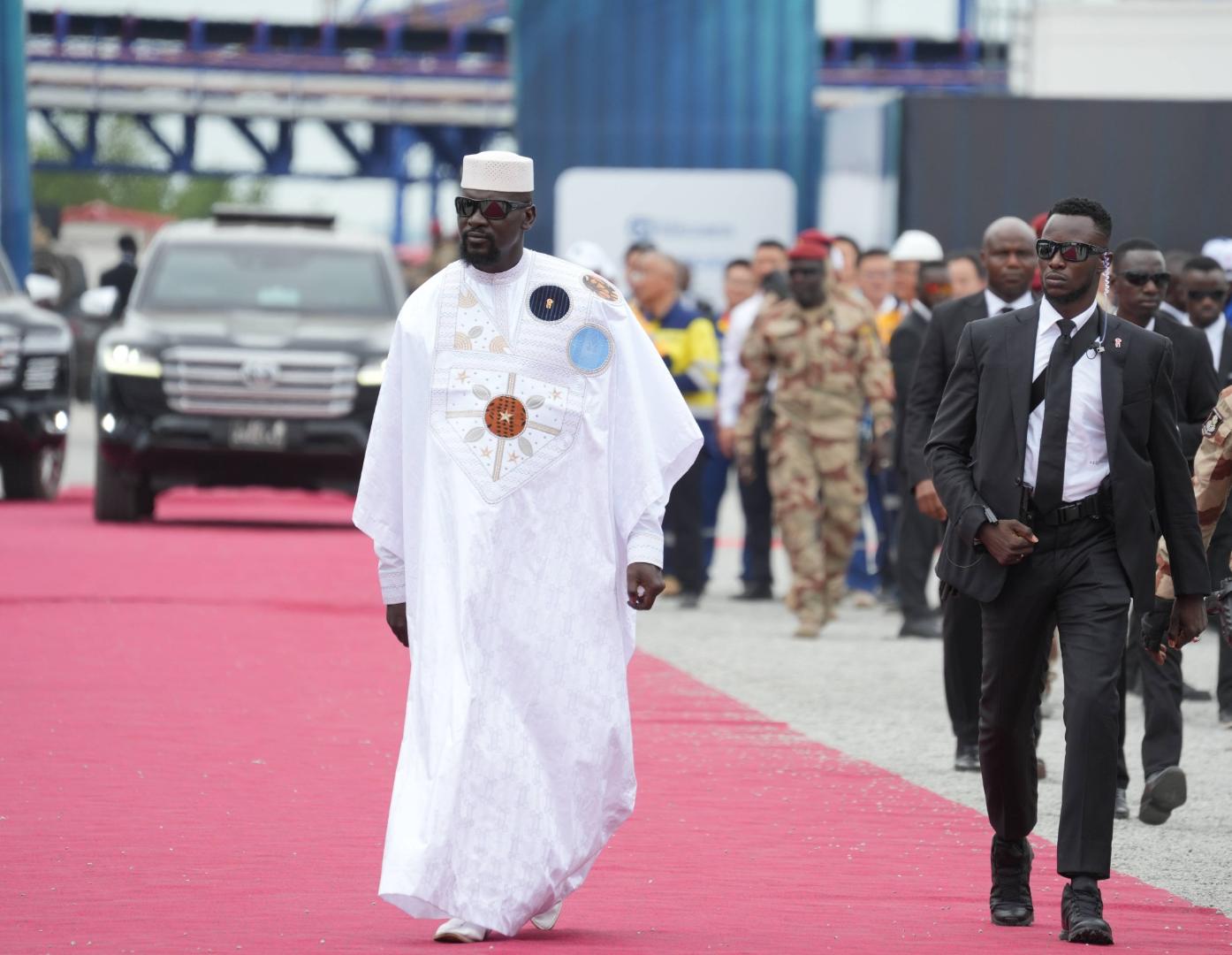 Mamady Doumbouya in traditioneller weißer Robe mit Stickereien schreitet auf einem roten Teppich, begleitet von mehreren Personen in Anzügen und Militäruniformen.