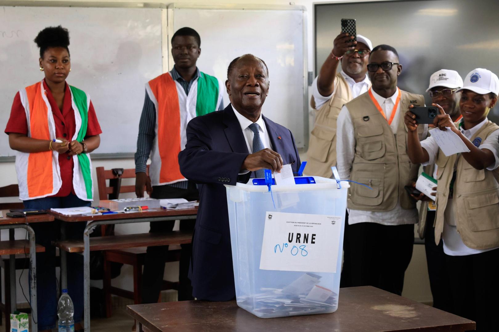 Ein Mann in dunklem Anzug wirft einen Wahlzettel in eine transparente Wahlurne mit der Aufschrift 'URNE', umgeben von mehreren Personen in orange-weißen und beige-weißen Westen, von denen einige Smartphones halten.