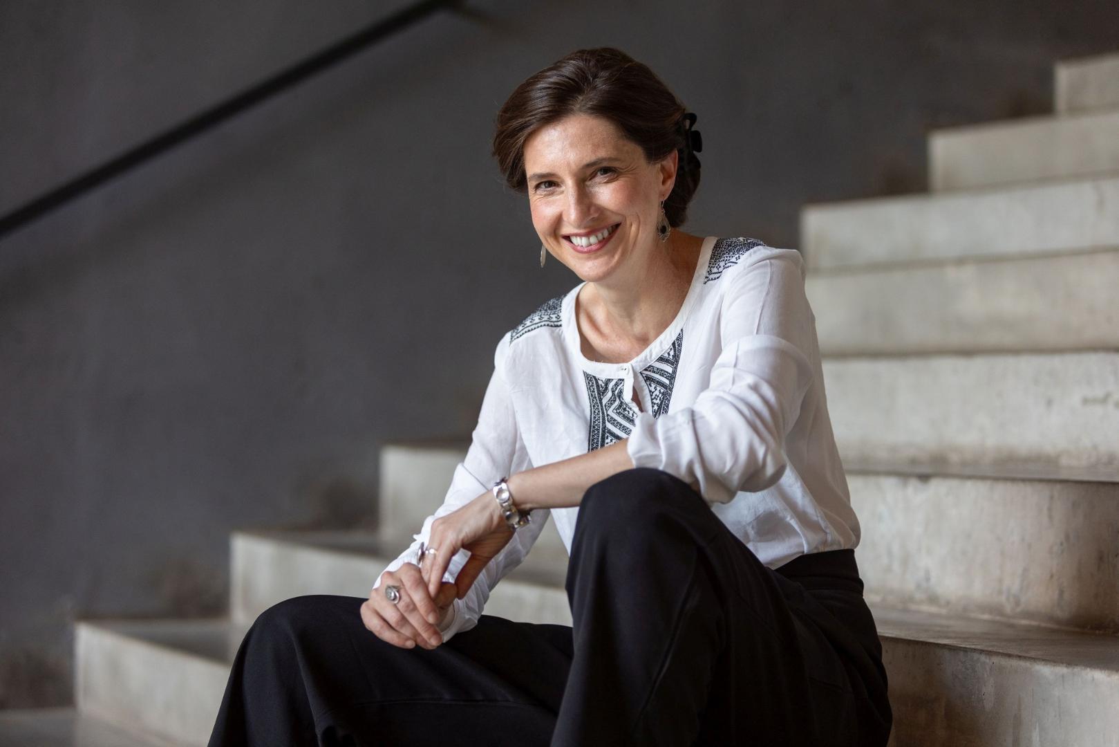 Daniela Danz sitzt auf einer Treppe, trägt eine weiße Bluse mit schwarzem Muster und schwarze Hosen, ihre Hände sind vor ihr verschränkt.
