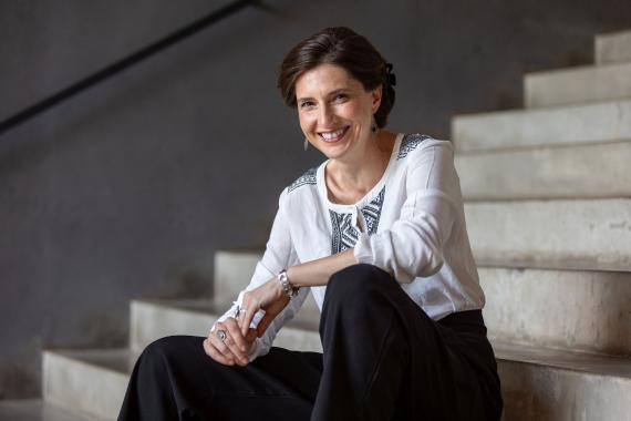 Daniela Danz sitzt auf einer Treppe, trägt eine weiße Bluse mit schwarzem Muster und schwarze Hosen, ihre Hände sind vor ihr verschränkt.