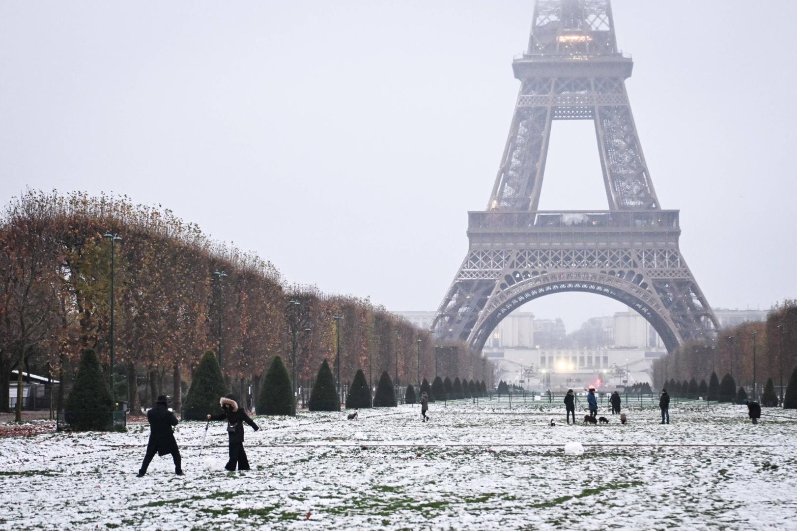 Der Eiffelturm steht im Hintergrund eines schneebedeckten Parks mit kahlen Bäumen und mehreren Personen, die sich im Vordergrund bewegen.