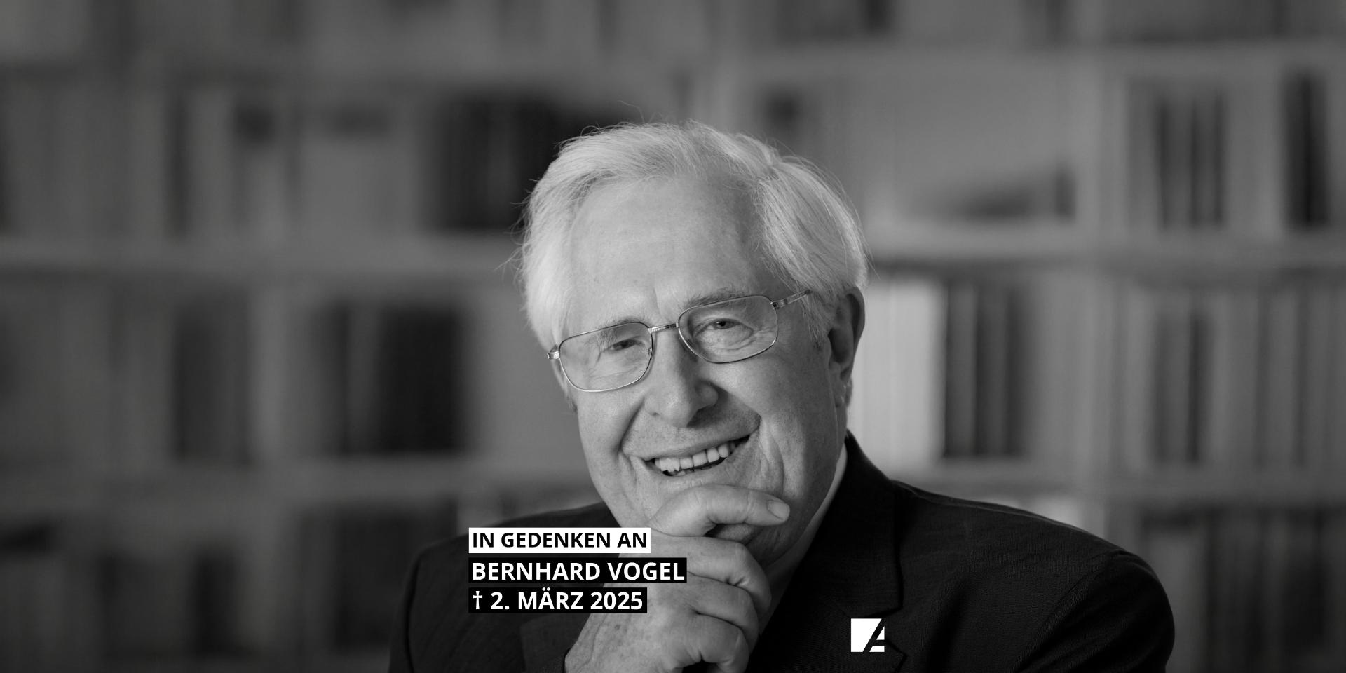 Mann mit weißem Haar in Anzug vor Bücherregal sitzend und die Hände vor dem Mund gefaltet