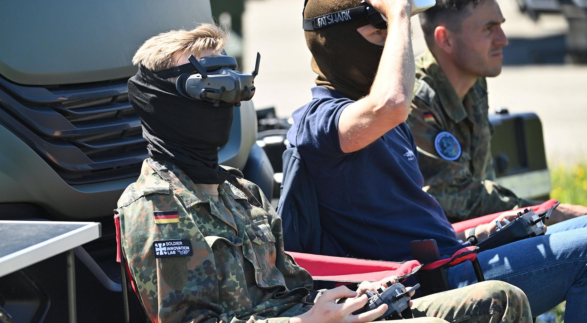 Zwei Personen mit schwarzer Gesichtsmaske sitzen auf Klappstühlen und steuern Drohnenfernbedienungen, eine trägt Bundeswehr-Uniform mit Abzeichen