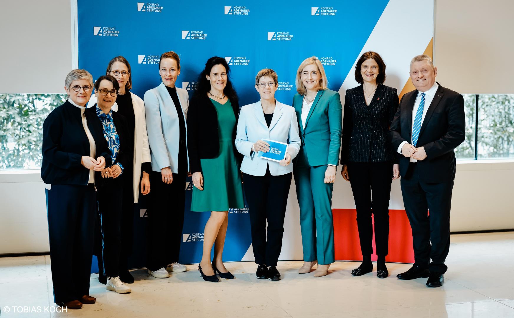Gruppe von acht Personen in formeller Kleidung vor blauem Hintergrund mit Logo der Konrad-Adenauer-Stiftung