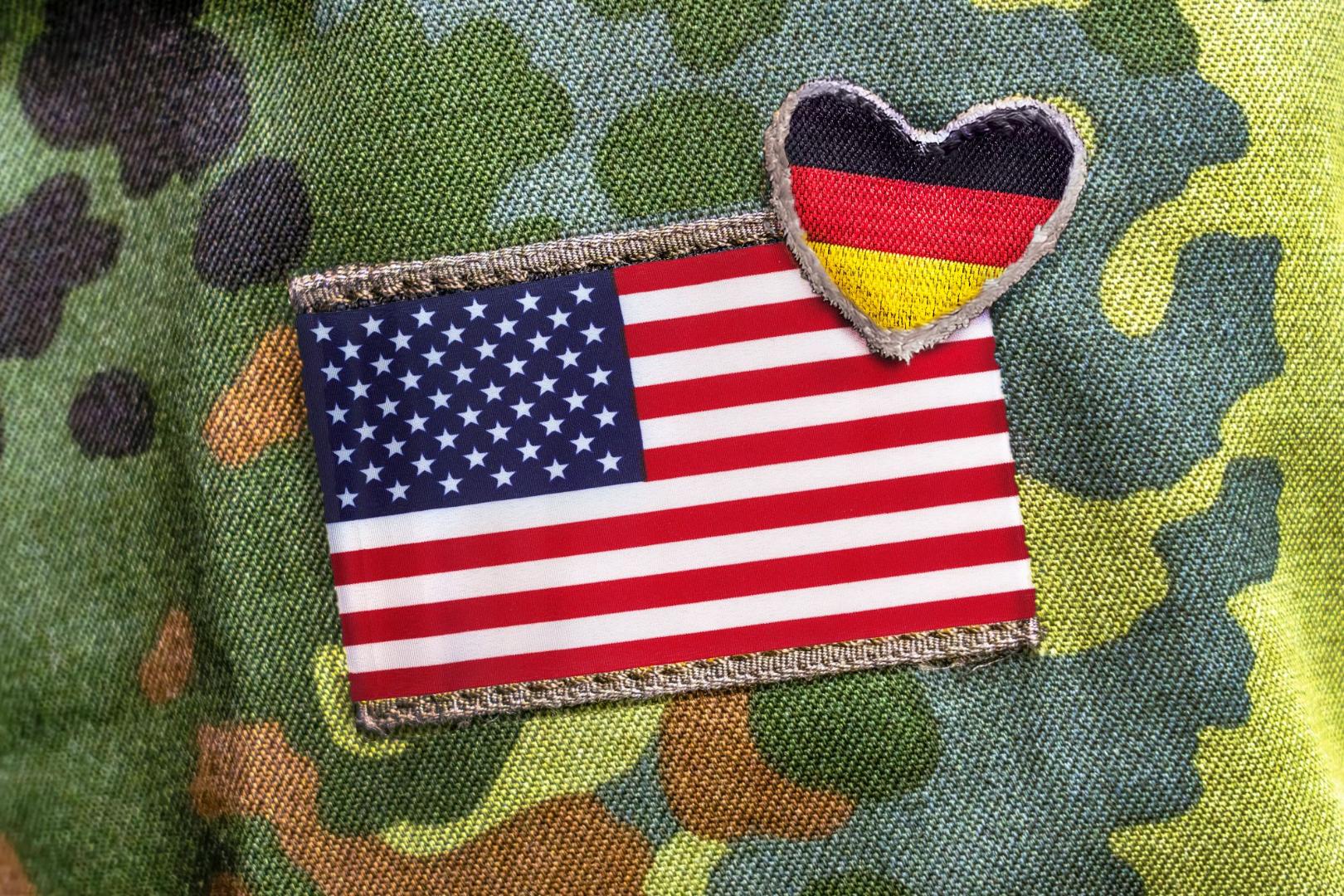 Auf einem Tarnmusterstoff sind zwei Stoffabzeichen befestigt, eine rechteckige US-amerikanische Flagge und ein herzförmiges Abzeichen mit der deutschen Flagge