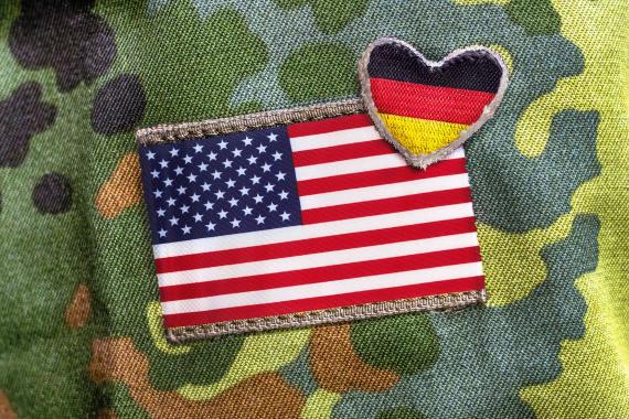 Auf einem Tarnmusterstoff sind zwei Stoffabzeichen befestigt, eine rechteckige US-amerikanische Flagge und ein herzförmiges Abzeichen mit der deutschen Flagge