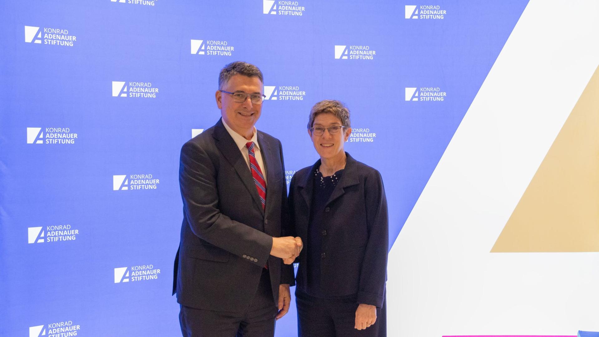 Zwei Personen in formeller Kleidung schütteln sich vor einer Wand mit dem Logo der Konrad-Adenauer-Stiftung