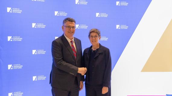 Zwei Personen in formeller Kleidung schütteln sich vor einer Wand mit dem Logo der Konrad-Adenauer-Stiftung