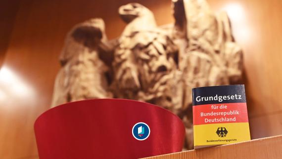 Vor dem Bundesadler im Sitzungssaal des Bundesverfassungsgericht steht eine Miniaturausgabe des Grundgesetz für die Bundesrepublik Deutschland sowie ein Barett eines Bundesverfassungsrichters.