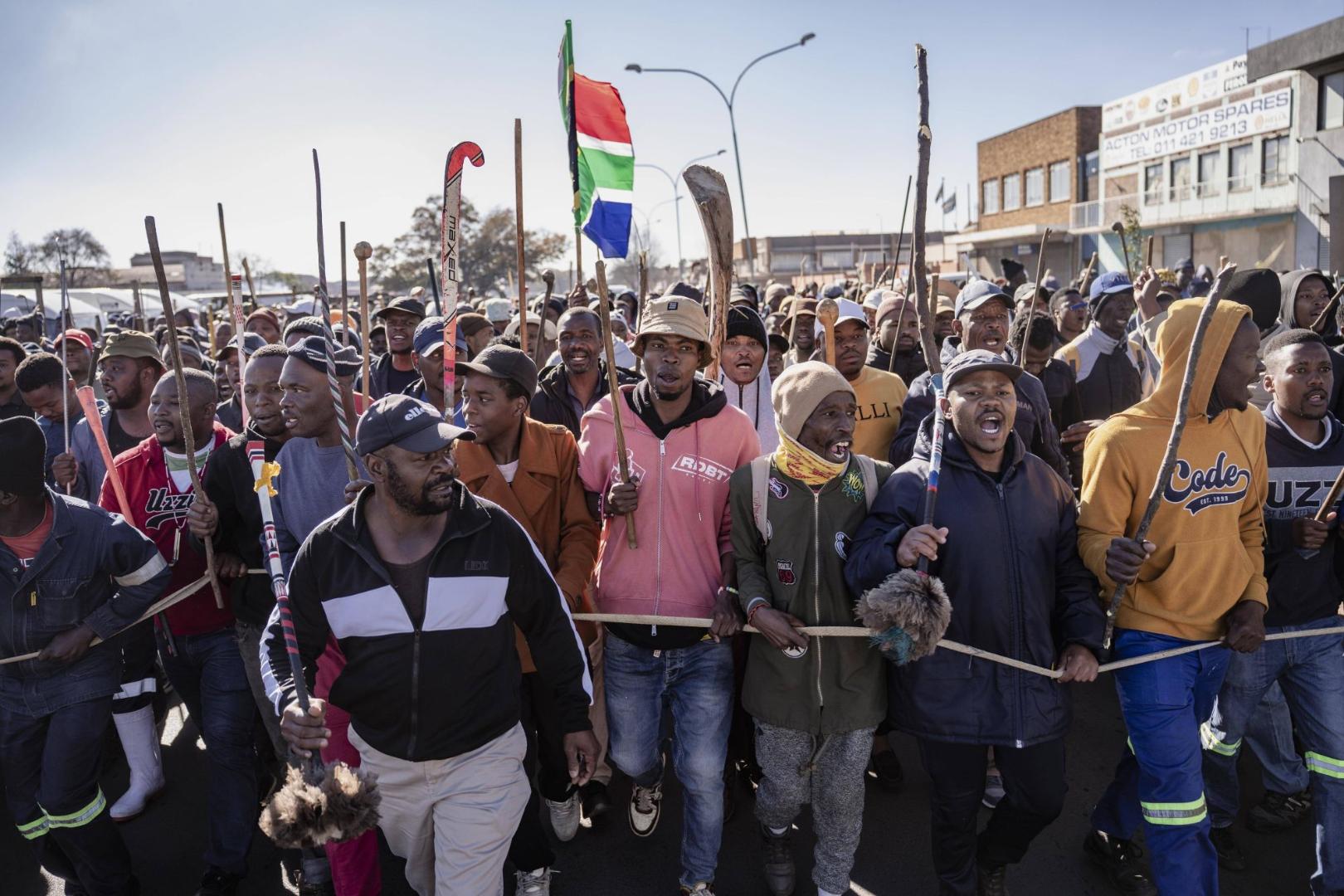 Gruppe von Menschen bei einer Demonstration in Südafrika, einige tragen Kapuzen und halten Stöcke, eine Person hält eine südafrikanische Flagge