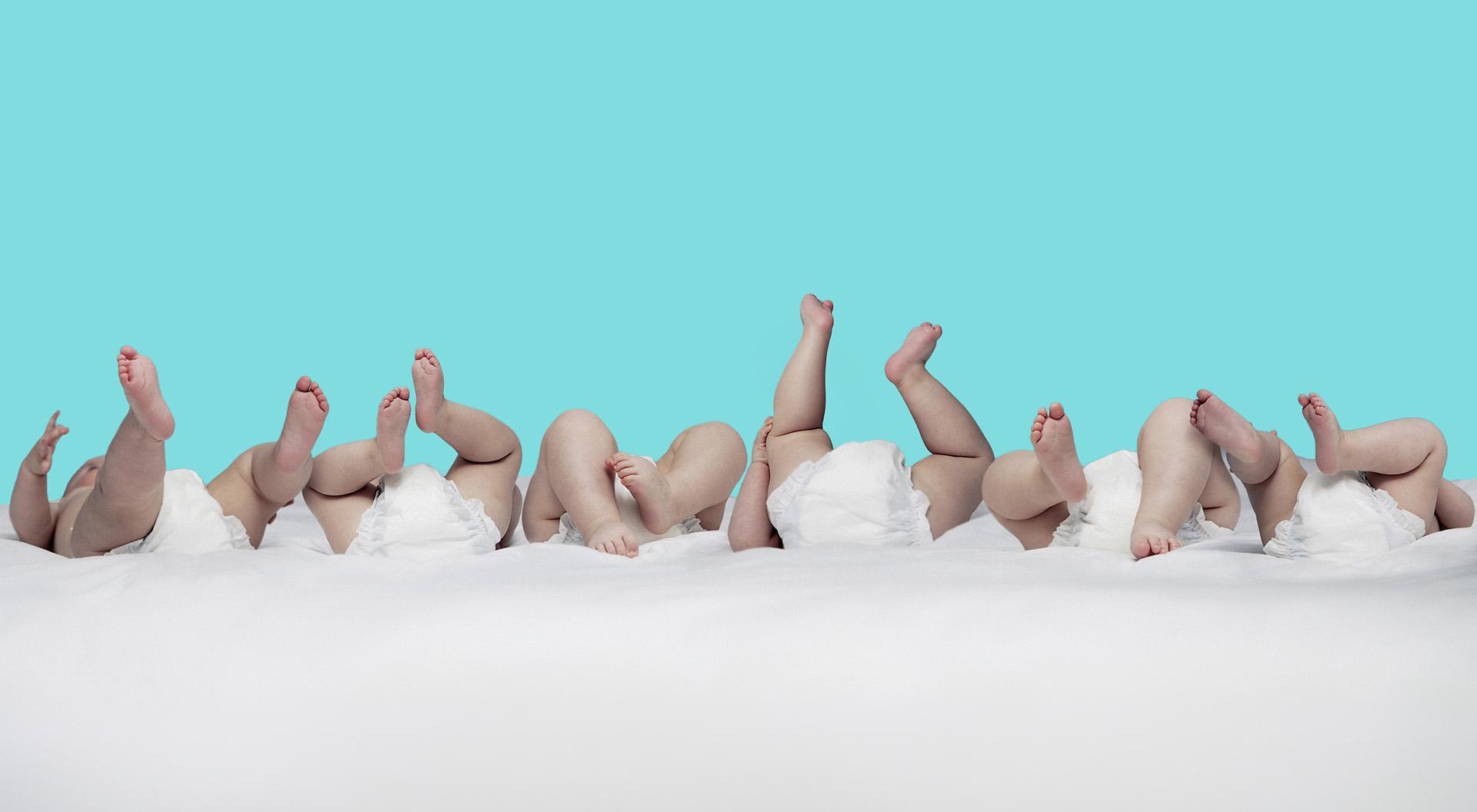 Mehrere Babys liegen nebeneinander mit hochgestreckten Beinen und weißen Windeln vor blauem Hintergrund