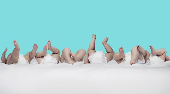Mehrere Babys liegen nebeneinander mit hochgestreckten Beinen und weißen Windeln vor blauem Hintergrund