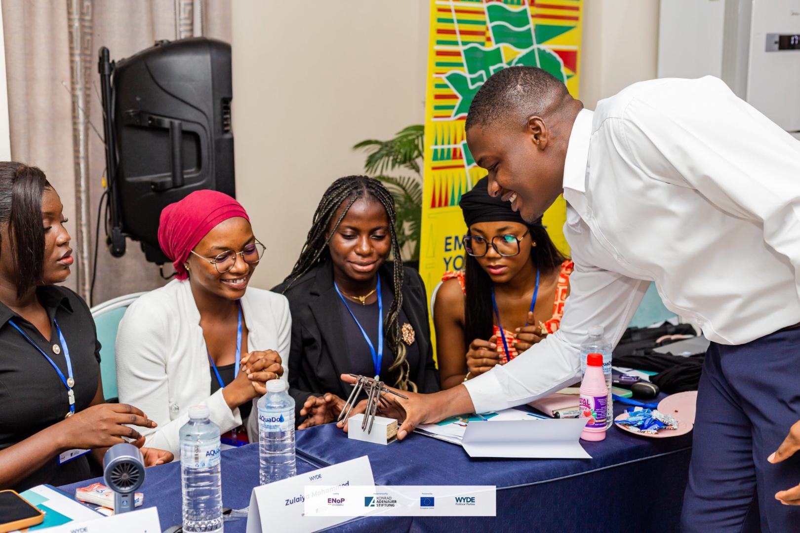 Five young adults gather around a table during a workshop. A man demonstrates a small technical model while the other participants watch attentively.