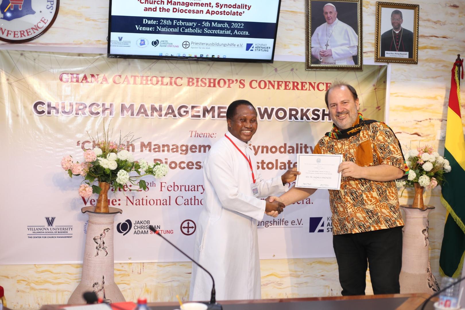 Two men shake hands and hold a certificate together at a church management workshop in Accra, Ghana, in front of a Ghana Catholic Bishops' Conference event banner.