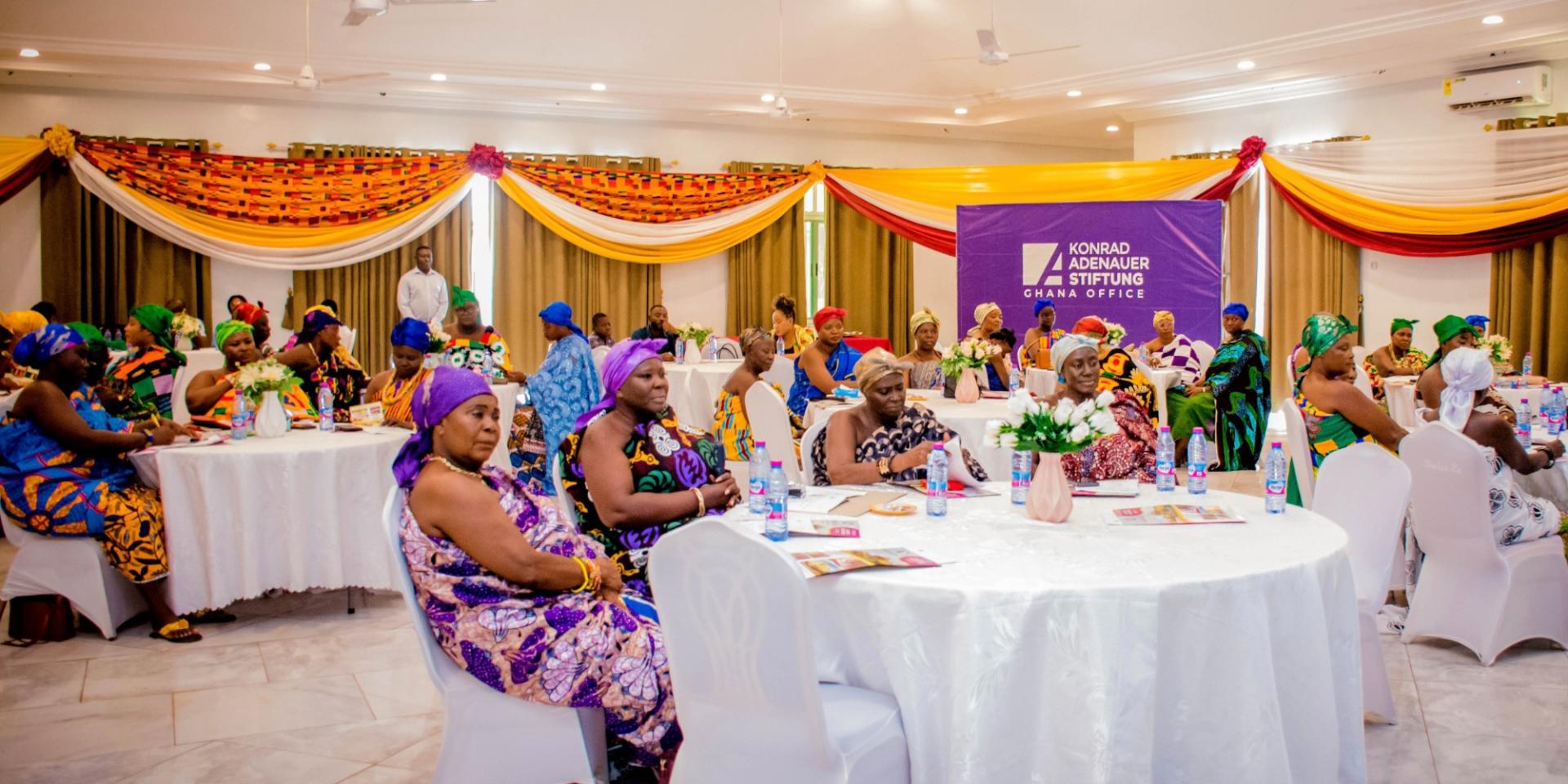 Women attending a Konrad Adenauer Stiftung workshop in Ghana sit at round tables wearing colorful traditional attire in a festively decorated conference hall.