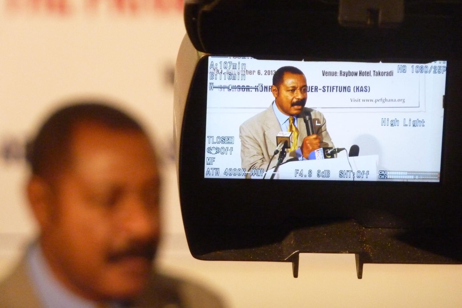 A man in a grey suit speaks into a microphone at a press conference in Takoradi, Ghana. He appears blurred in the foreground, while the camera display shows his speech in focus.