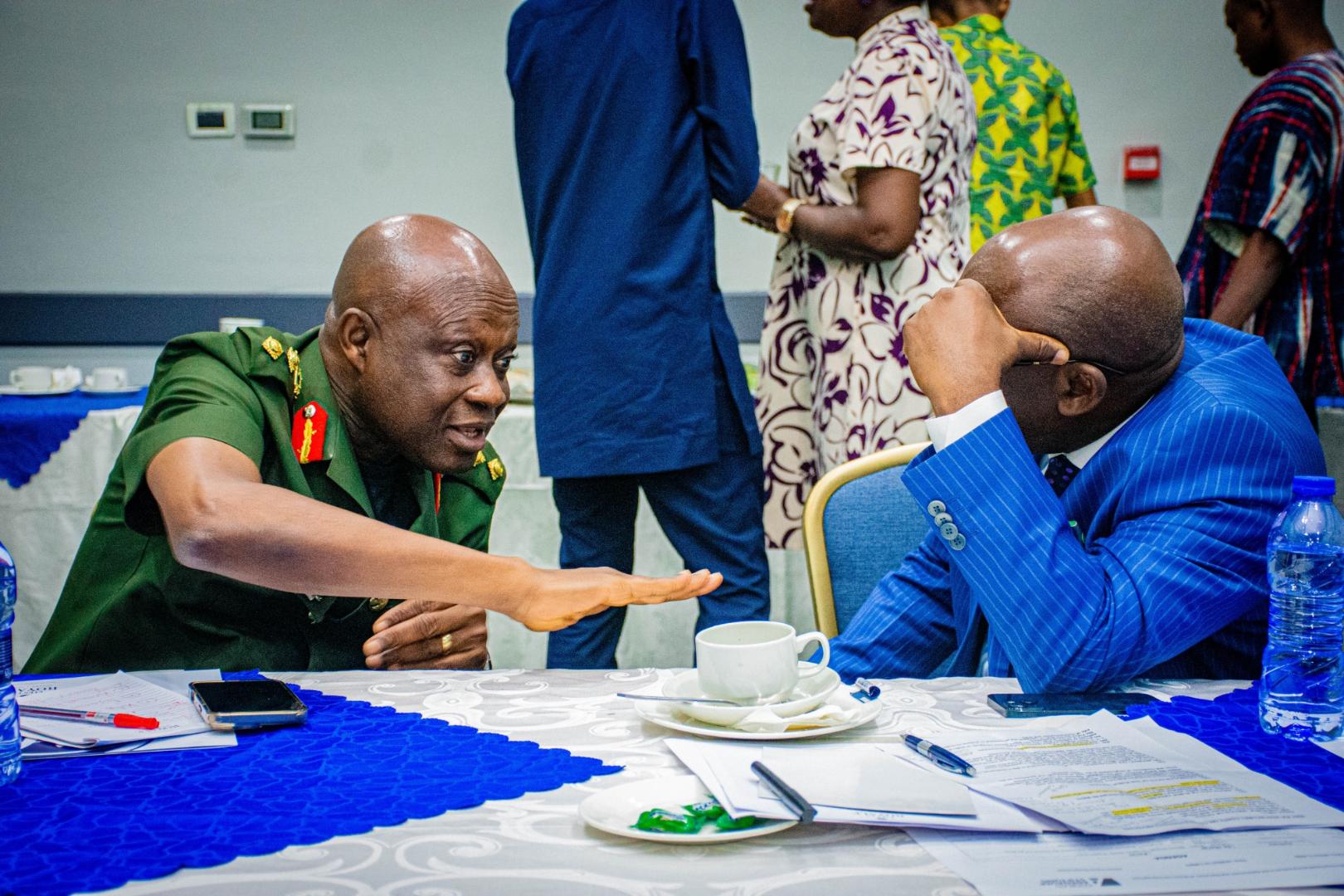 A man in a green military uniform gestures while speaking with a man in a blue suit during a workshop. Both are seated at a conference table, while other participants converse in the background.