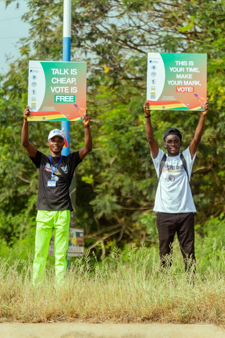 Two young men stand outdoors in Ghana holding posters encouraging voter participation. The posters display the messages 