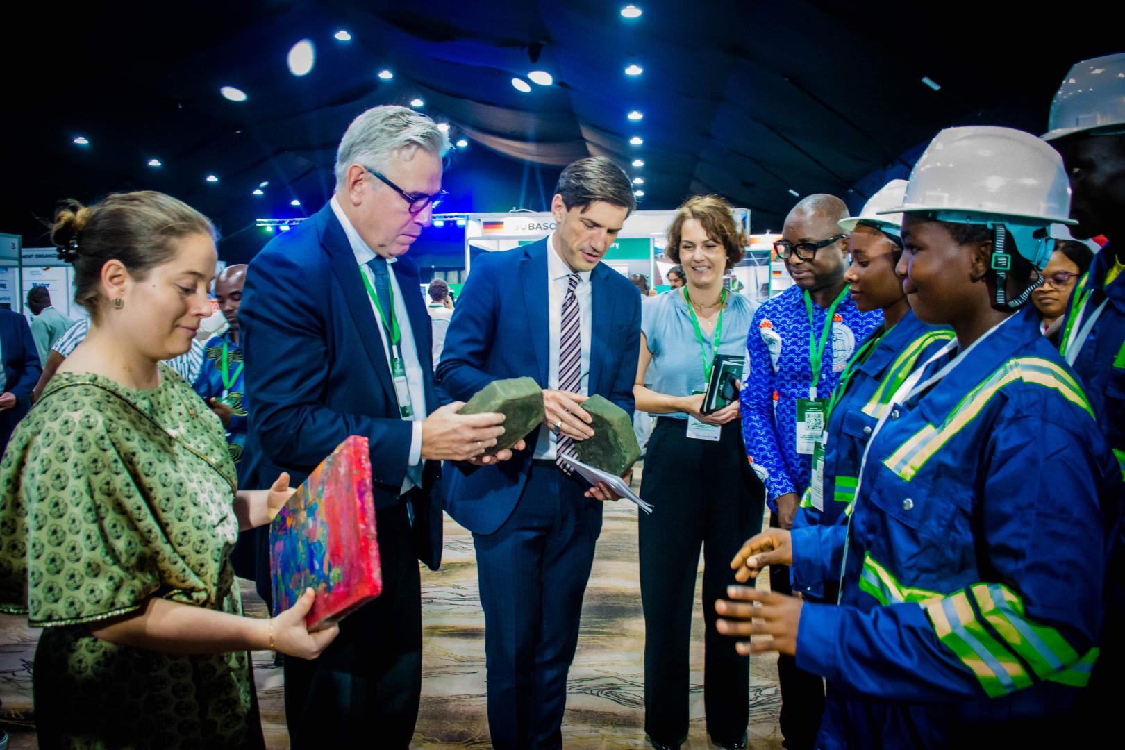 Participants at the West African Clean Energy and Environment Trade Fair (WACEE) examine various material samples at an exhibition stand. Two women in blue workwear and safety helmets explain the displayed products.