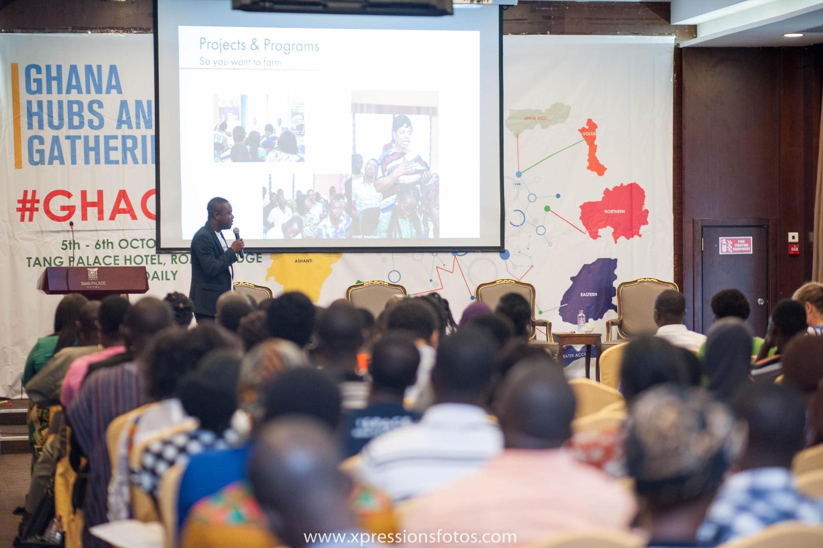A speaker presents agricultural projects and programmes at a forum on entrepreneurship and innovation in Ghana. Numerous participants follow the presentation in a conference hall.