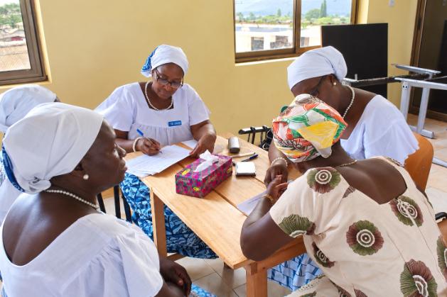 Four women sit around a wooden table in a bright room. Two women dressed in white with headscarves talk with two other women, while one of them takes notes on a sheet of paper.