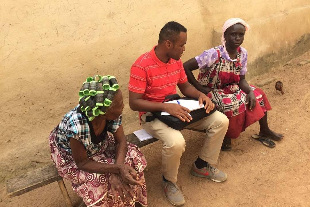 A man in a red polo shirt sits with two women on a wooden bench in front of a yellow wall. He holds a notepad and pen and turns toward one of the women. Both women wear patterned clothing.